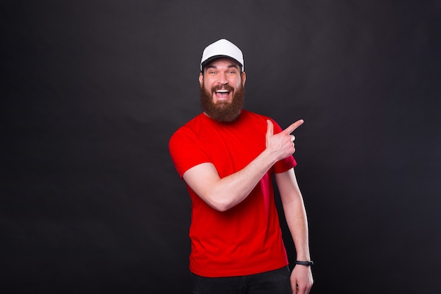 Portret van verbaasde jonge man met baard in rood t-shirt wijzend op hoek over zwart