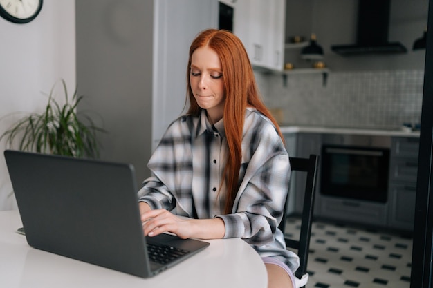 Portret van verbaasde jonge vrouw typend op laptopcomputer zittend aan tafel peinzend op zoek naar scherm in keuken Aantrekkelijke vrouwelijke student leren op afstand