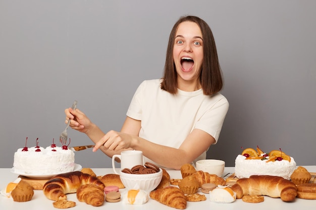 Portret van verbaasde vrolijke aantrekkelijke vrouw met bruin haar zittend aan tafel geïsoleerd over grijze achtergrond eten cake kijken camera schreeuwen geluk breken dieet