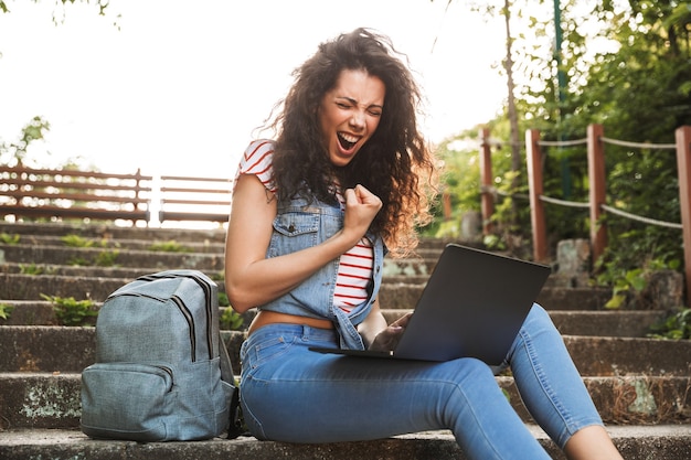 Portret van verbijsterde gelukkige vrouw balde vuisten en schreeuwen, zittend op de trap in het park op een zonnige dag, en zilveren laptop op knieën te houden