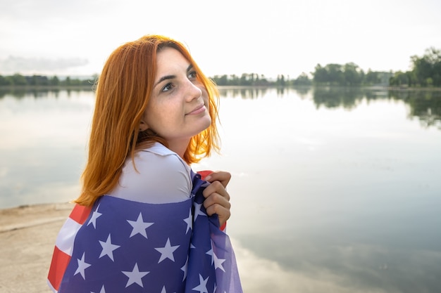 Portret van verdrietig roodharig meisje met de nationale vlag van de VS op haar schouders. Jonge vrouw die de onafhankelijkheidsdag van de Verenigde Staten viert.