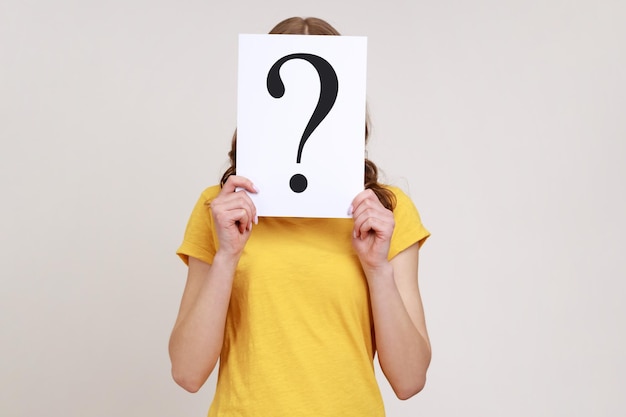 Portret van verlegen vrouw in gele casual Tshirt haar gezicht achter Witboek met vraagteken verbergen slimme oplossing vragen om advies Indoor studio shot geïsoleerd op grijze achtergrond