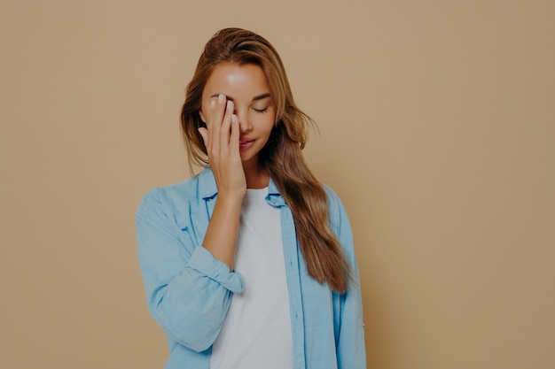 Portret van vermoeide en geërgerde jonge vrouw die ogen verbergt omdat ze zich schaamt en gezichtspalmgebaar maakt, hoofd naar beneden kantelend, boos en geïrriteerd over beige achtergrond in blauw oversized shirt