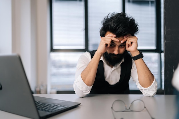 Portret van vermoeide indiase zakenvrouw in stress werkt op laptop terwijl ze aan kantoortafel bij raam zit en hand vasthoudt aan slapen migraineaanval overwerkte man bezorgd over problemen op het werk