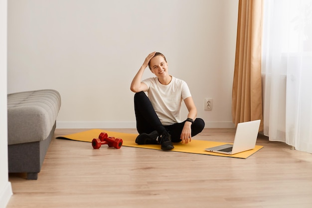 Portret van vermoeide jonge volwassen vrouw zittend op yogamat met blije gezichtsuitdrukking na online training met trainer vrouw met sportief lichaam met wit t-shirt en leggins