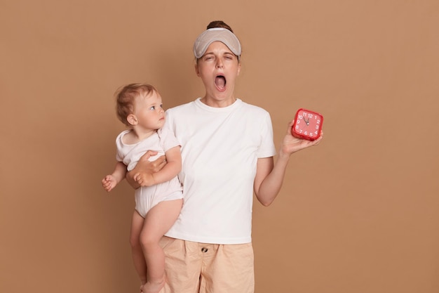 Portret van vermoeide slaperige vrouw met wit t-shirt en blinddoek geeuwen met wijd open mond met baby dochter en rode wekker tijd om wakker te worden poseren geïsoleerd over bruine achtergrond