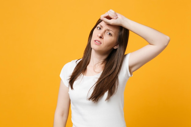 Portret van vermoeide uitgeputte jonge vrouw in witte vrijetijdskleding op zoek naar camera, hand op hoofd geïsoleerd op geeloranje muurachtergrond in studio. Mensen levensstijl concept. Bespotten kopie ruimte.