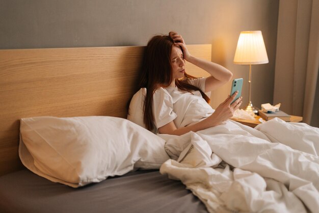 Portret van vermoeide zieke roodharige vrouw die online videogesprek heeft via smartphone die thuis in bed ligt Zieke roodharige vrouw die mobiele telefoon gebruikt om met dokter te praten
