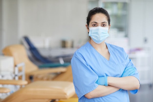 Portret van verpleegster in masker en in uniform status met haar gekruiste armen en in het ziekenhuis