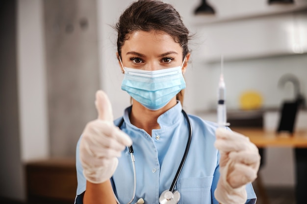 Portret van verpleegster met gezichtsmasker op om thuis te zitten, spuit met vaccin vast te houden en duimen opdagen tijdens het coronavirus.