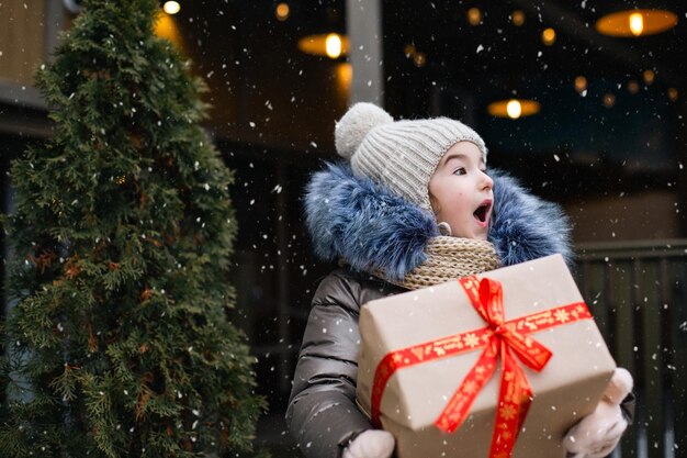 Portret van verrast meisje met een geschenkdoos voor Kerstmis op een stadsstraat in de winter met sneeuw op een feestelijke markt met versieringen en verlichting. Warme kleding, gebreide muts, sjaal en bont. Ruimte kopiëren