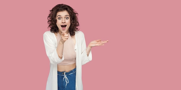 Portret van verrast mooie brunette jonge vrouw met krullend kapsel in casual stijl staan, wijzen, verbaasd en kijken naar de camera. indoor studio-opname geïsoleerd op roze achtergrond kopie ruimte.