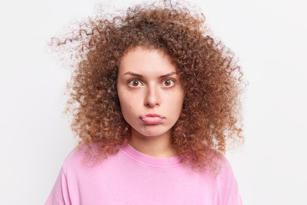 Foto portret van verrast tienermeisje met krullend borstelig haar portemonnees lippen heeft verbijsterd uitdrukking ziet er gefascineerd gekleed in casual kleding poses tegen witte muur. reactie concept