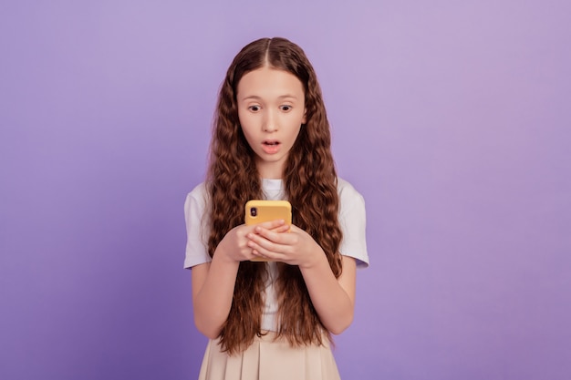 Portret van verrast verbaasd meisje houdt telefoon open mond post onverwachte reactie op paarse achtergrond