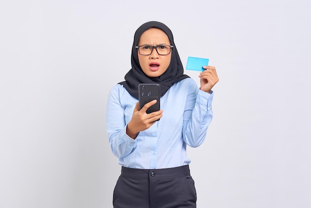 Portret van verraste jonge Aziatische vrouw die mobiele telefoon houdt en creditcard toont die op witte achtergrond wordt geïsoleerd