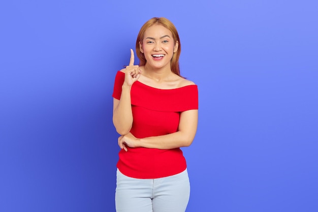 Portret van verraste jonge Aziatische vrouw in rode jurk en blond haar, wijzend met idee geïsoleerd op paarse achtergrond