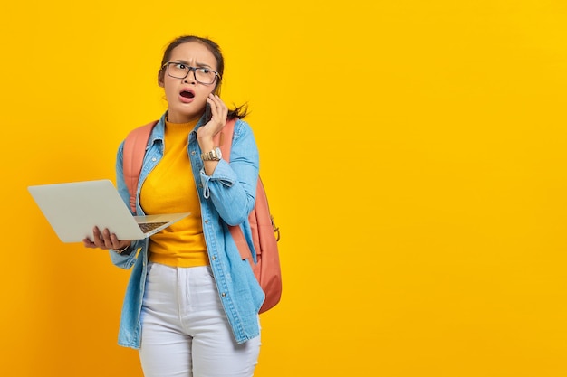 Portret van verraste jonge Aziatische vrouw student in casual kleding met rugzak praten op mobiele telefoon en laptop geïsoleerd op gele achtergrond te houden. Onderwijs in universiteitsconcept