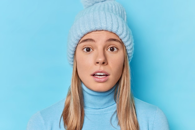 Foto portret van verraste jonge europese vrouw hapt naar adem van verwondering kan niet geloven in schokkend nieuws draagt winter gebreide muts en casual coltrui geïsoleerd over blauwe achtergrond. menselijke reacties concept