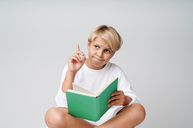 Portret van verwarde blonde jongen poseren geïsoleerd over grijze muur leesboek wijzen.