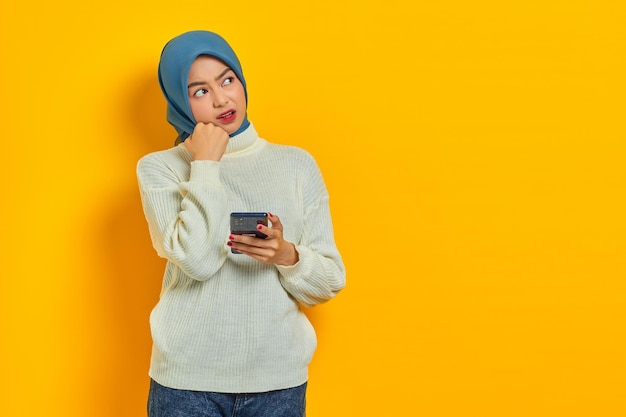 Portret van verwarde jonge Aziatische vrouw in witte trui en hijab met behulp van mobiele telefoon en kijkt serieus na over een vraag geïsoleerd over gele achtergrond Mensen islam religieus concept