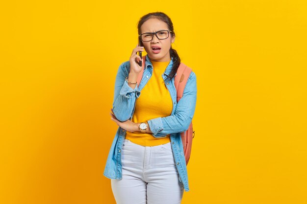 Portret van verwarde jonge Aziatische vrouw student in casual kleding met rugzak praten op smartphone en kijken naar camera geïsoleerd op gele achtergrond. Onderwijs in universiteitsconcept