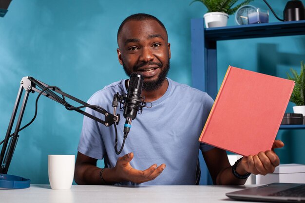 Portret van vlogger met audiomicrofoon die podcast opneemt over het verhaal in rood mockupboek in vlogstudio. Contentmaker zit aan bureau met nieuwe roman voor video op sociale media.