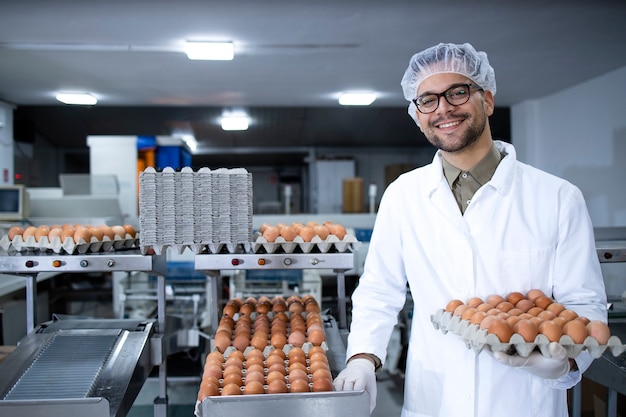 Portret van voedsel fabrieksarbeider met haarnetje en hygiënische handschoenen die eieren houden door industriële transport- en verpakkingsmachine in voedselverwerkingsfabriek.