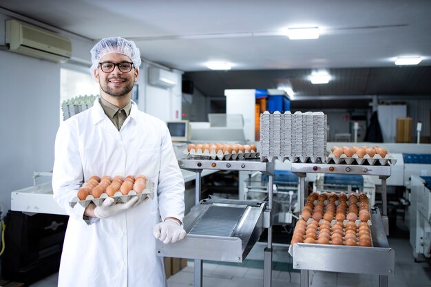 Portret van voedsel fabrieksarbeider met haarnetje en hygiënische handschoenen die eieren houden door industriële transport- en verpakkingsmachine in voedselverwerkingsfabriek.