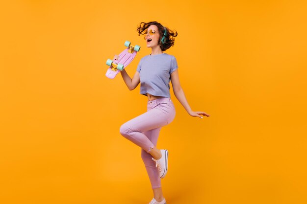 Portret van volledige lengte van gelukkig Kaukasisch vrouwelijk model met roze longboard Studiofoto van mooi krullend meisje dat muziek luistert in hoofdtelefoons op oranje achtergrond