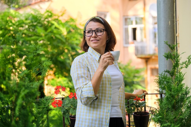 Portret van volwassen huisvrouw vrouw, vrouw met kopje koffie op open balkon versierd met groene planten