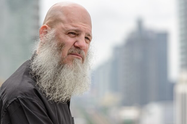 Portret van volwassen kale hipster man met lange baard tegen uitzicht over de stad buitenshuis