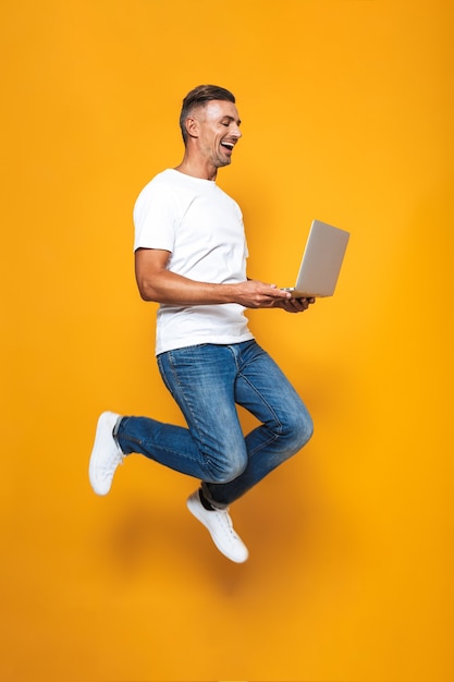 Portret van volwassen man 30s in wit t-shirt houden en gebruiken van zilveren laptop geïsoleerd op geel