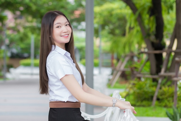 Portret van volwassen Thaise student in universitair studentenuniform. Aziatisch mooi meisje zit gelukkig glimlachend aan de universiteit in de buitenlucht met een achtergrond van tuinbomen.