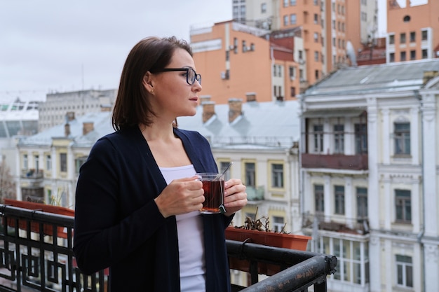 Portret van volwassen vrouw met kopje thee staande op buitenbalkon