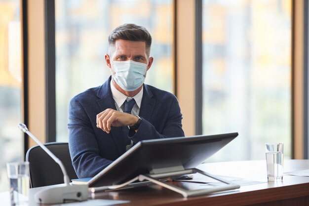 Portret van volwassen zakenman in beschermend masker kijken zittend op conferentie