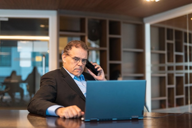 Portret van volwassen zakenman met behulp van laptopcomputer en mobiele telefoon op kantoor