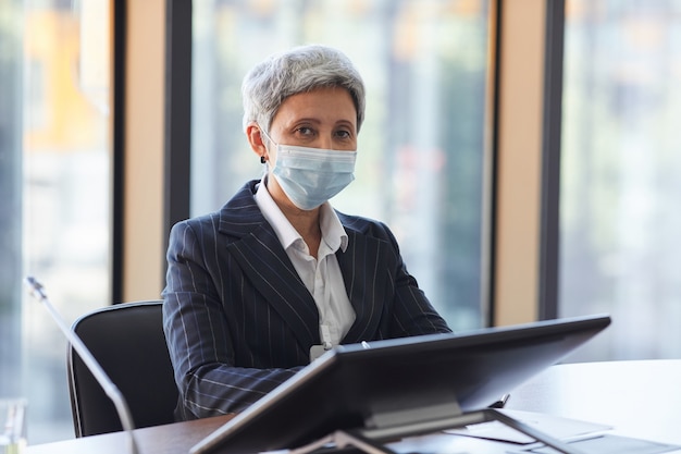 Portret van volwassen zakenvrouw in beschermend masker zitten aan de tafel met computer en kantoor kijken