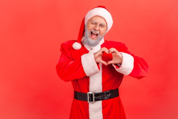 Portret van vriendelijke positieve kerstman in bril met grijze baard hart gebaar met handen tonen en knipogen naar camera liefde verzenden op vakantie Indoor studio shot geïsoleerd op rode achtergrond