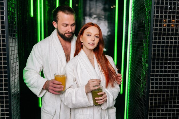 Portret van vrij jong verliefd stel in badjas met glas met vers geperst vitaminesap in handen staande knuffelend kijkend naar camera genietend van ontspanning in spa salon