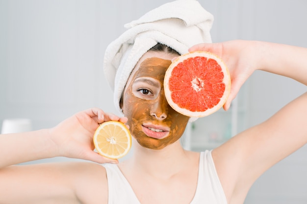 Portret van vrij jonge vrouw met kleimasker op haar gezicht met plakjes citroen en grapefruit, gezichtsverzorging