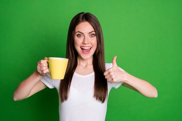 Portret van vrij positief opgewonden brunette meisje die cacao drinkt, toont duim omhoog geïsoleerd over groene kleur achtergrond