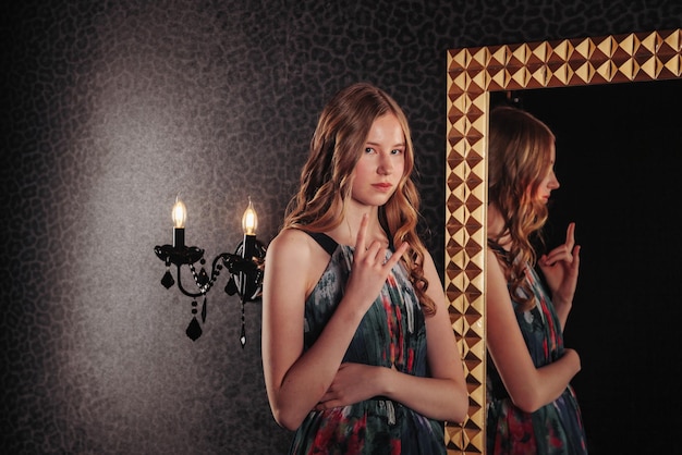 Portret van vrij schattig tienermeisje in een elegante jurk bij spiegel in donkere stijlvolle interieur van woonkamer kinderen emoties en poseren Concept van stijl mode en schoonheid Kopieer ruimte voor site