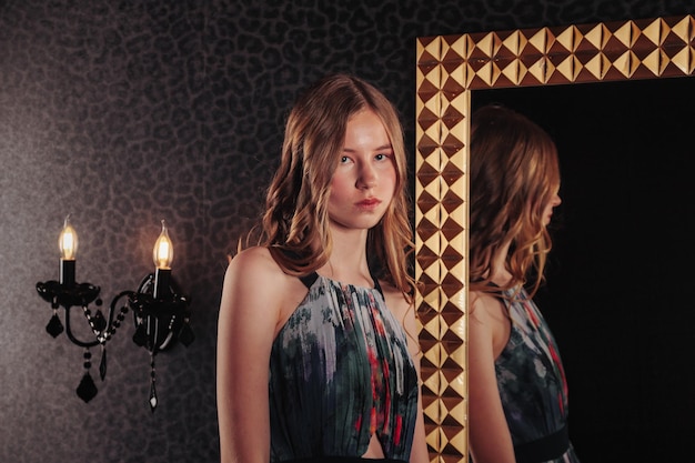 Portret van vrij schattig tienermeisje in een elegante jurk bij spiegel in donkere stijlvolle interieur van woonkamer kinderen emoties en poseren Concept van stijl mode en schoonheid Kopieer ruimte voor site