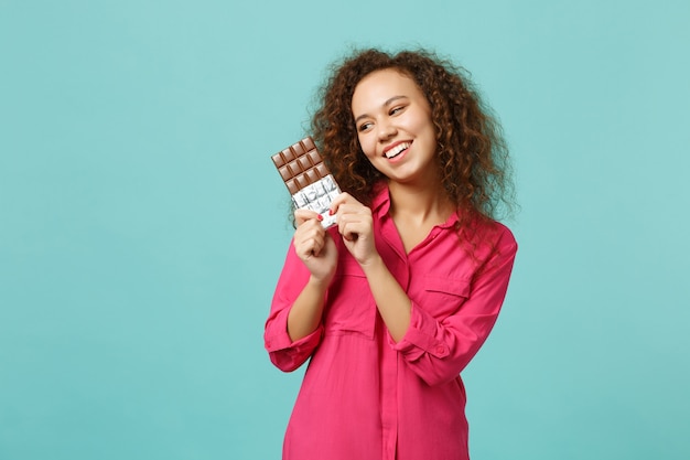 Portret van vrolijk Afrikaans meisje in casual kleding met in de hand chocoladereep geïsoleerd op blauwe turquoise muur achtergrond in studio. Mensen oprechte emoties, lifestyle concept. Bespotten kopie ruimte.