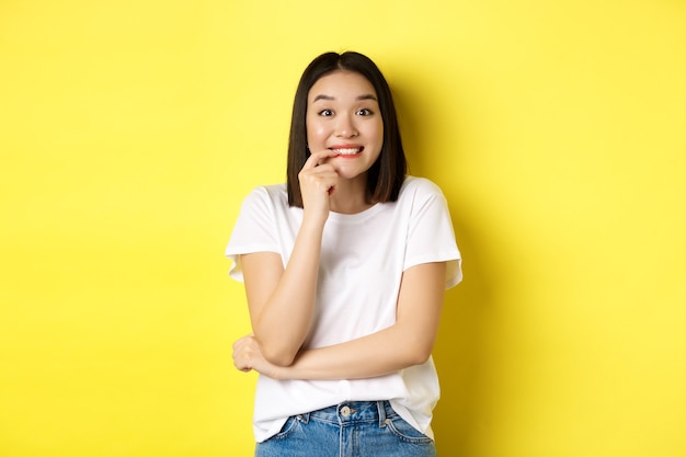 Portret van vrolijk Aziatisch meisje wil iets, kijkend met verlangen en verleiding naar de camera, lachend naar de camera, staand ovr geel.