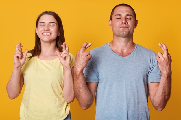 Portret van vrolijk energiek paar dat zich dicht bij elkaar bevindt, hun vingers kruist, armen opheft, handen sluit, wensen maakt, nadenkt over positieve dingen, bidden. familie concept.