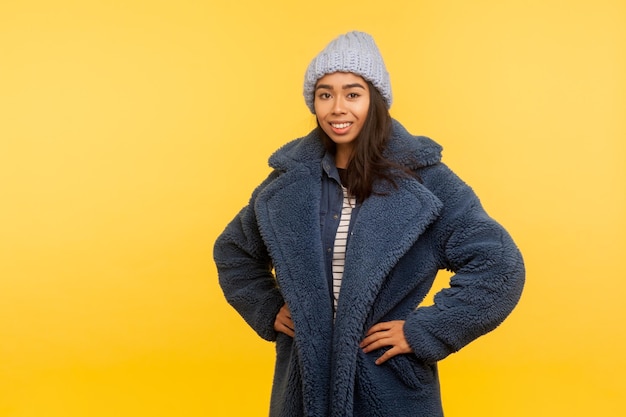 Portret van vrolijk gelukkig meisje dragen warme winter muts en bontjas glimlachend vreugdevol glamour fashion model in stijlvolle stedelijke outfit poseren voor camera binnen studio opname geïsoleerd op gele achtergrond