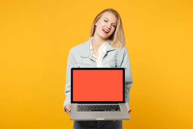 Portret van vrolijk jong meisje in denim casual kleding met laptop pc-computer met leeg leeg scherm geïsoleerd op geeloranje achtergrond in studio. Mensen levensstijl concept. Bespotten kopie ruimte.