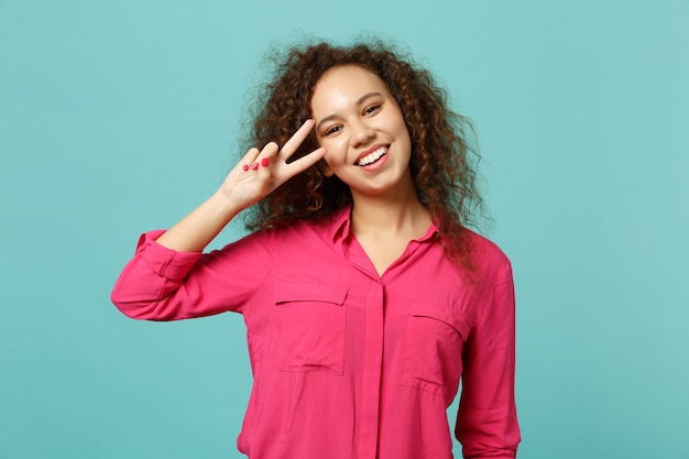 Portret van vrolijk lachend Afrikaans meisje in casual kleding met overwinningsteken geïsoleerd op blauwe turquoise muur achtergrond in studio. Mensen oprechte emoties, lifestyle concept. Bespotten kopie ruimte.