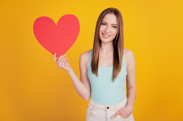 Portret van vrolijke aanhankelijke dame houdt liefdessymbool rood hartvorm ansichtkaart op gele achtergrond
